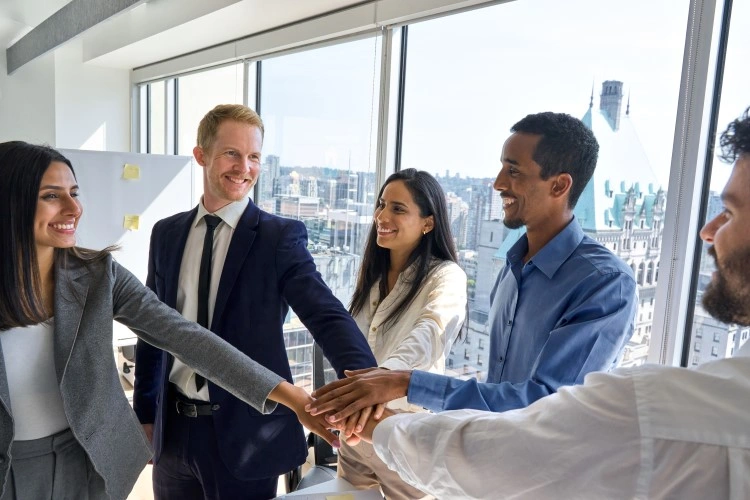 Smiling coworkers do “hands-in” during a meeting in the glassed-in room.