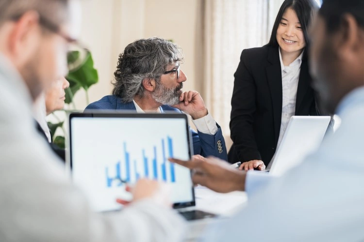 Business meeting: in the foreground a laptop with a bar chart and a pointing hand, in the background a conversation of colleagues.