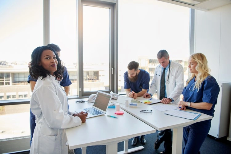Bright hospital room - doctors and nurses work on reports, one person looks at the camera.