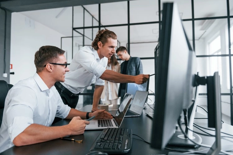 IT consultation in a modern office — a man shows something on the screen, a colleague works on a laptop.