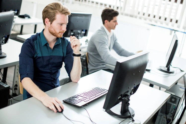 Open space/IT lab: two people focused on desktop work, keyboard and mouse in the foreground.
