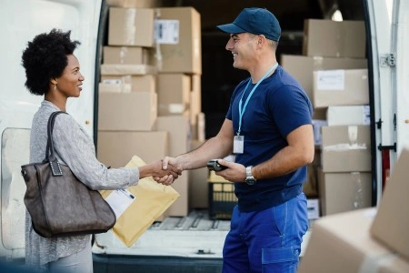 The courier gives the parcel to the customer in front of the delivery car; in the background, boxes with packages.