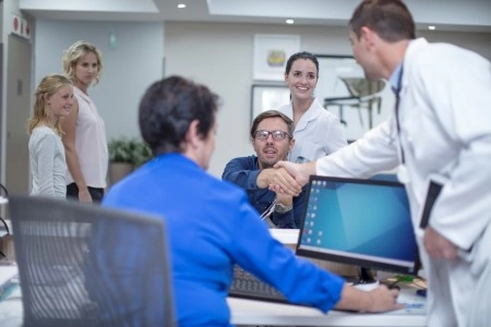 A doctor in a white coat greets the patient with a handshake at the reception; in the background, medical staff and a computer stand.