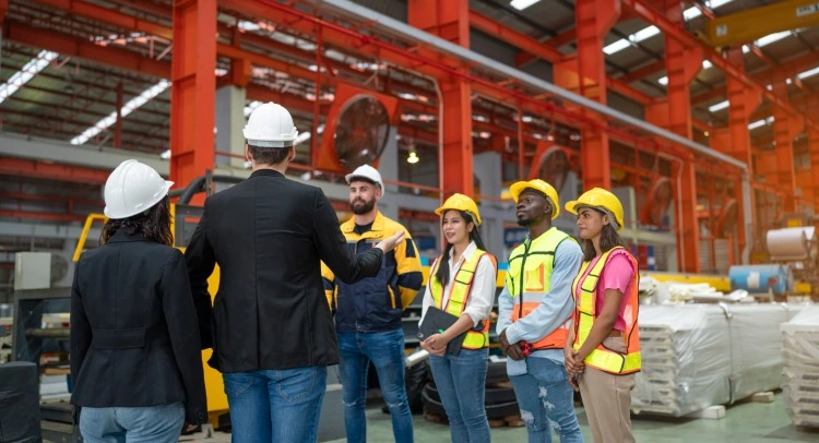 Production hall: a supervisor in a helmet conducts a health and safety briefing with a group of workers in reflective vests.