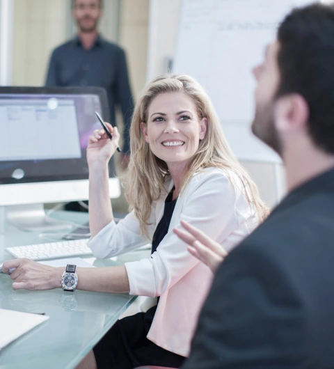Smiling woman talking to co-worker at desk in modern office; monitor and whiteboard in background.
