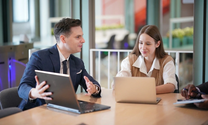 Meeting at the table: a man in a suit gestures as he discusses something with a woman; both have their laptops open.