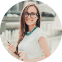 A smiling woman with glasses, wearing a white shirt and a blue necklace, stands in front of a fountain, holding a pen and a notebook. The lighting is soft and natural.
