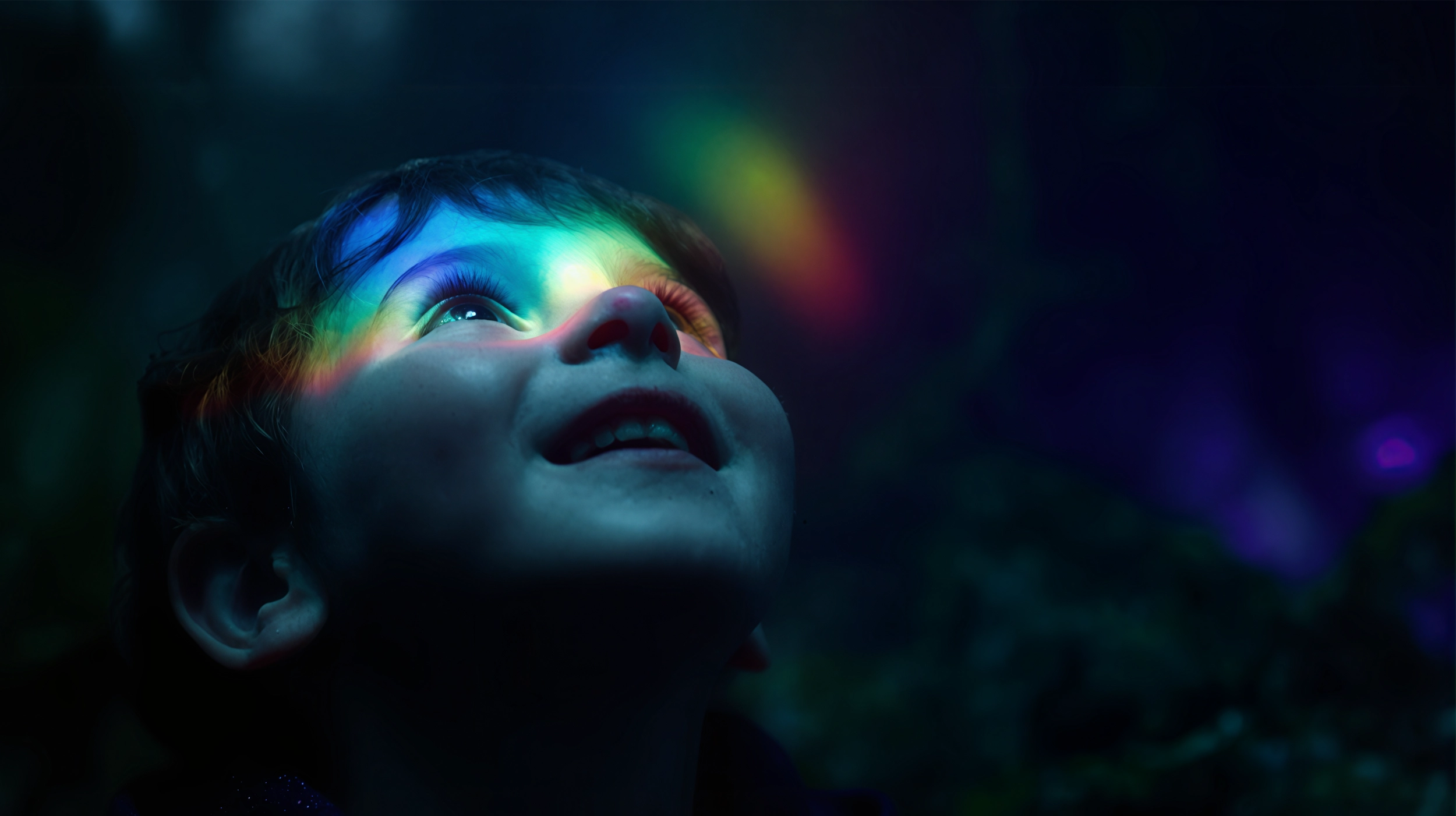 Young child looking up with a colorful light spectrum reflected on their face against a dark background.