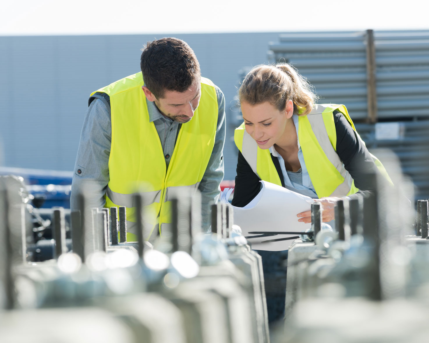Two construction workers in reflective vests examining blueprints at a construction site with concrete pillars.