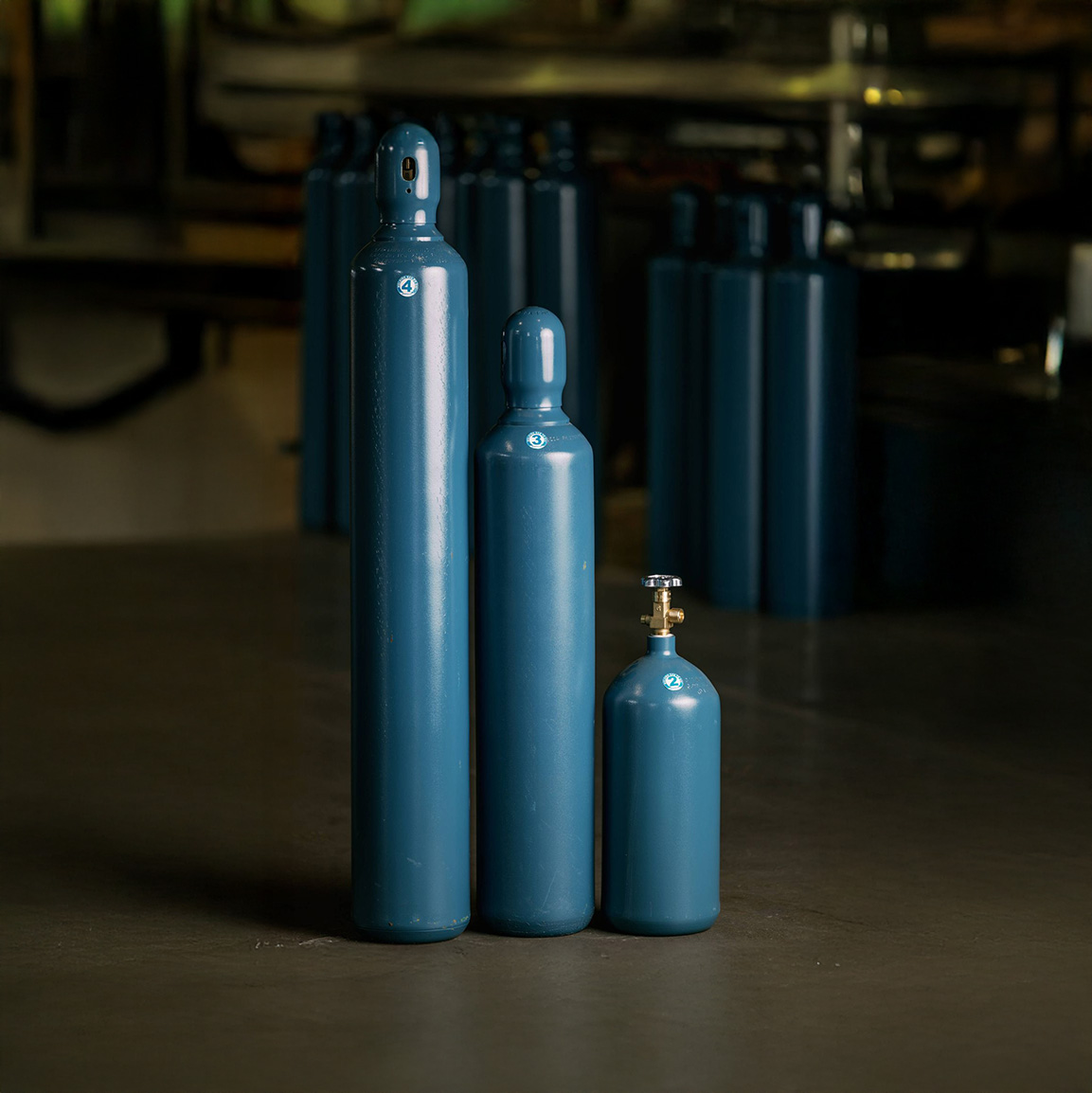 Three blue industrial gas cylinders of varying sizes standing upright on a concrete floor in a dimly lit room.