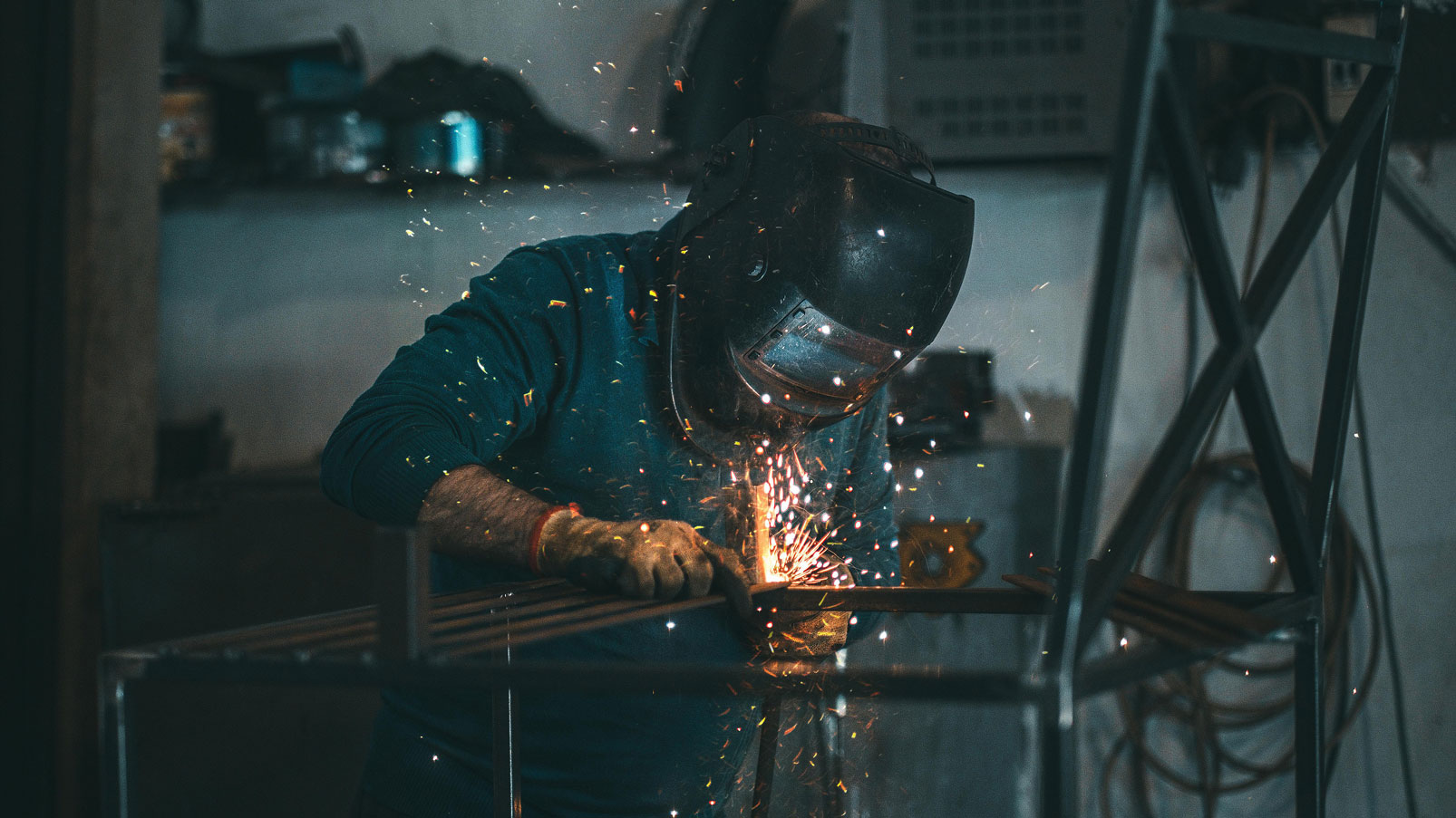 A person wearing a welding helmet and gloves welding metal with sparks flying in a workshop.