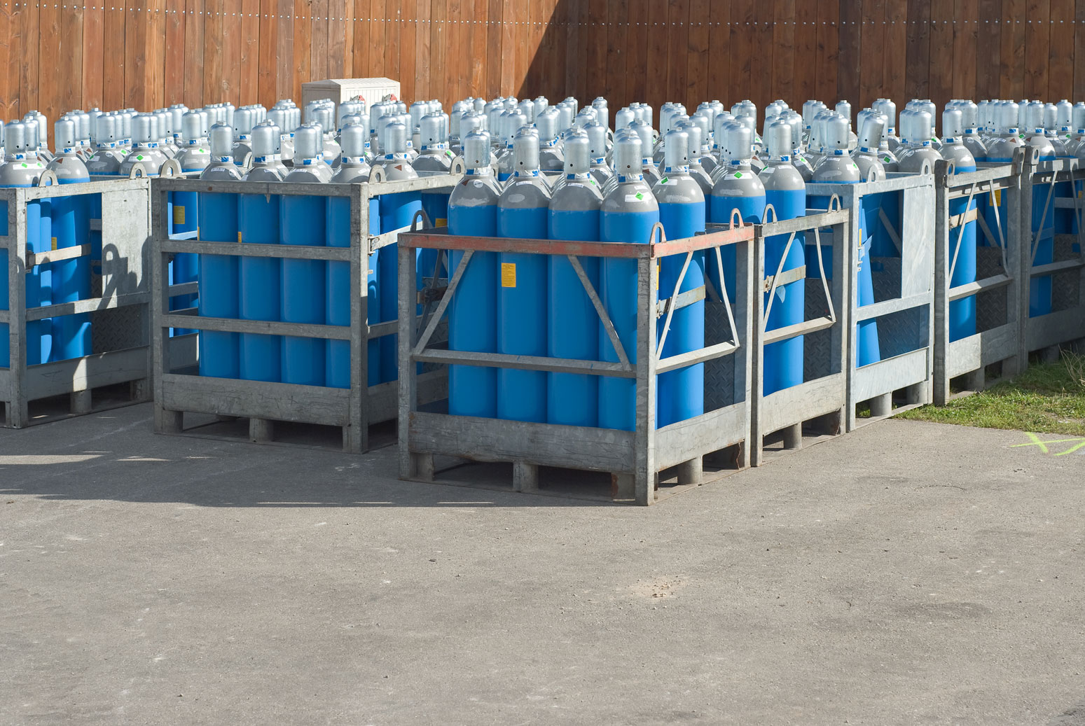 Multiple metal crates holding grouped blue gas cylinders with gray tops arranged outdoors on pavement near a wooden fence.