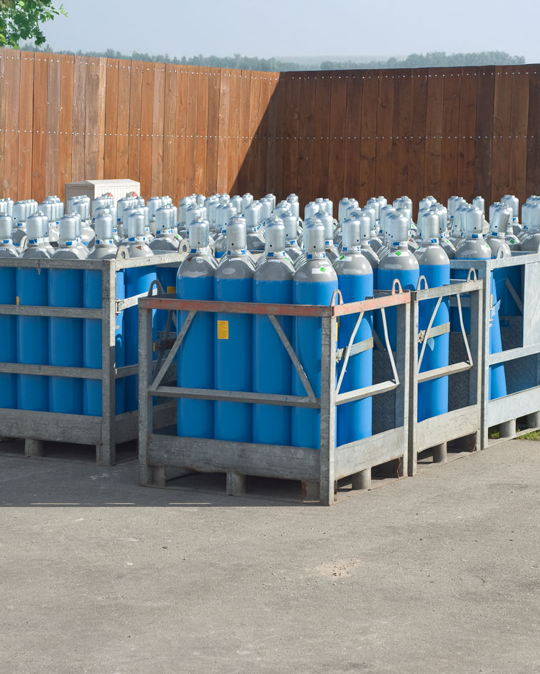 Metal crates filled with multiple blue compressed gas cylinders with silver tops, placed on a paved surface beside a wooden fence.