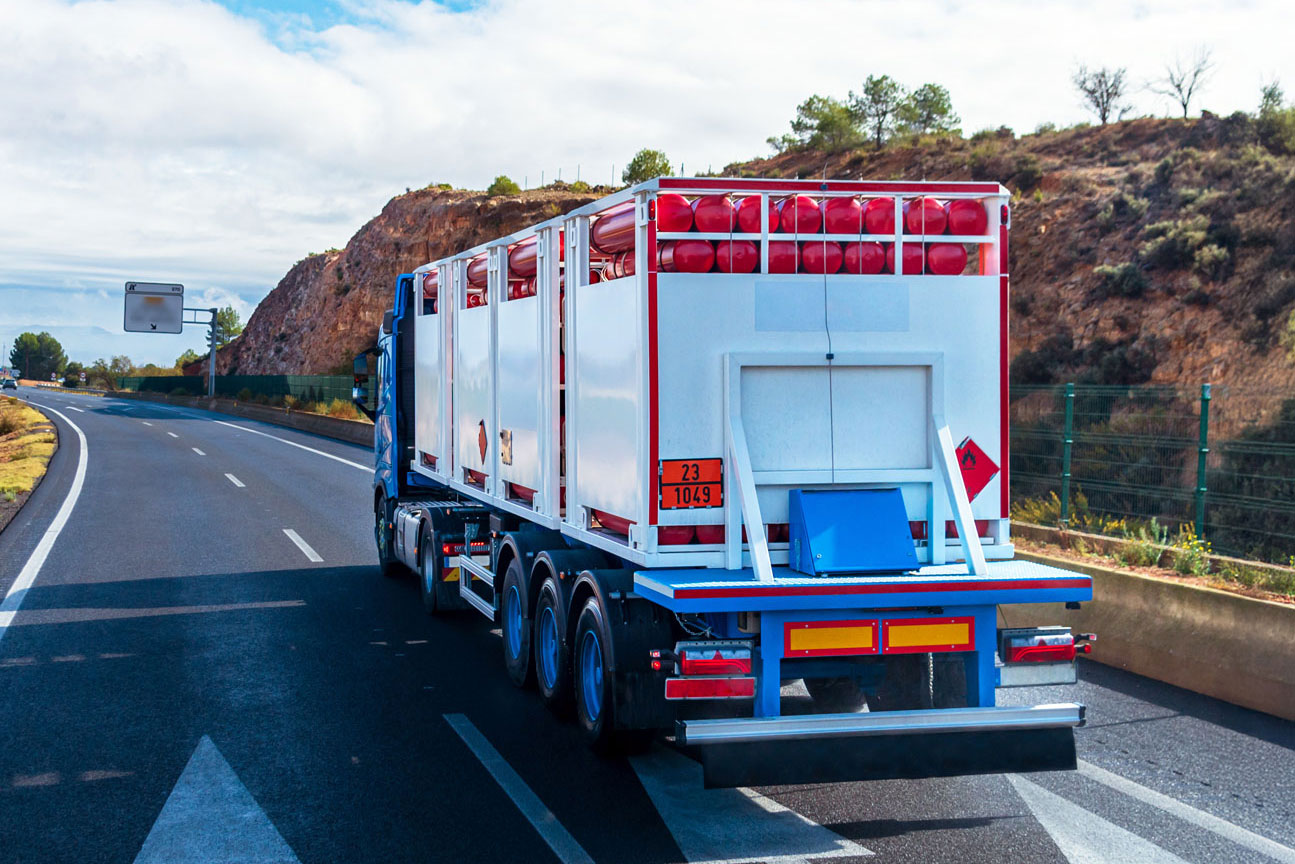 Blue truck carrying red gas cylinders driving on a highway with rocky hills in the background.
