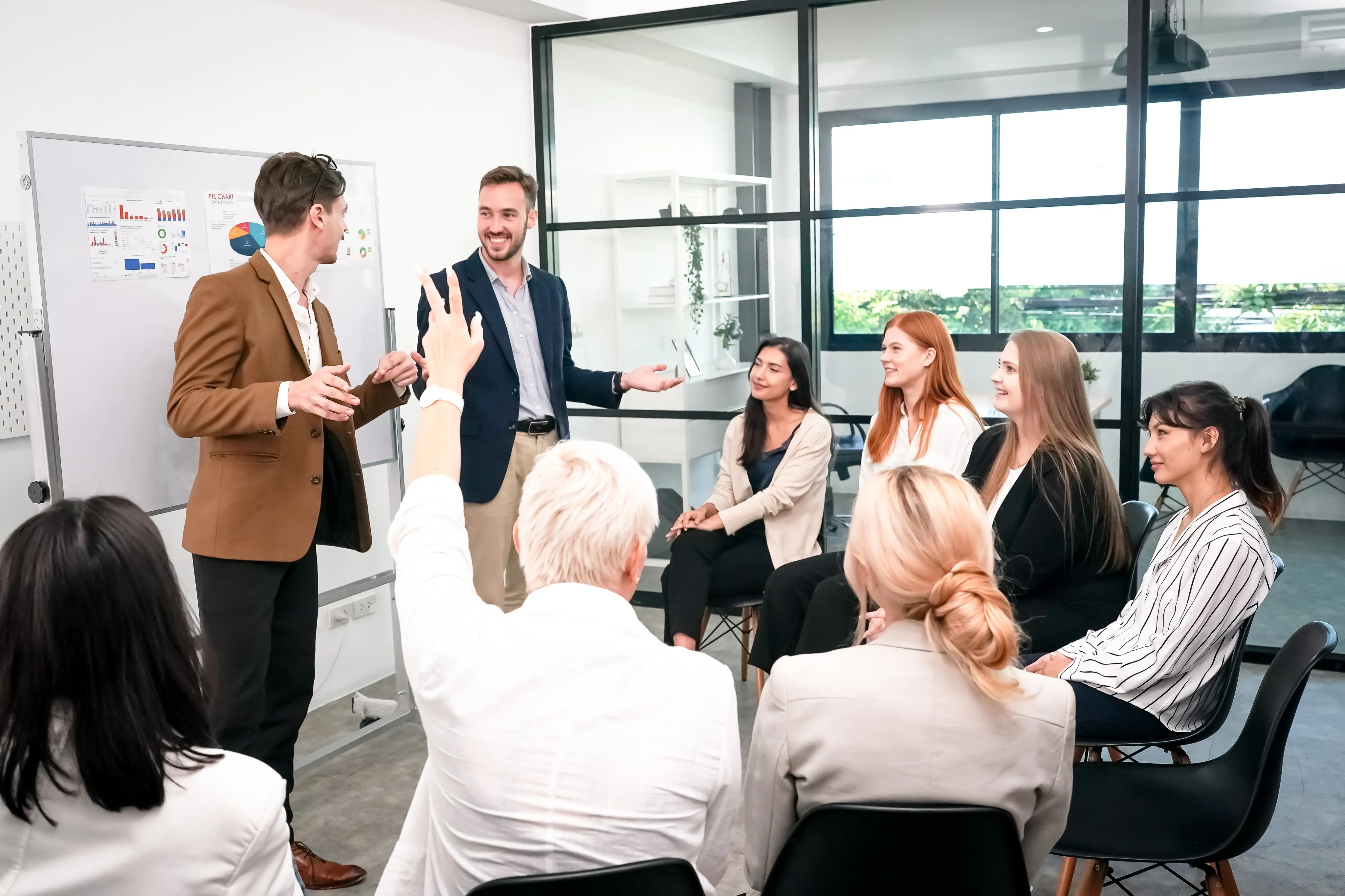 A group of professionals sitting in a modern office while two men stand near a whiteboard with charts, engaging in discussion; one person raises a hand.