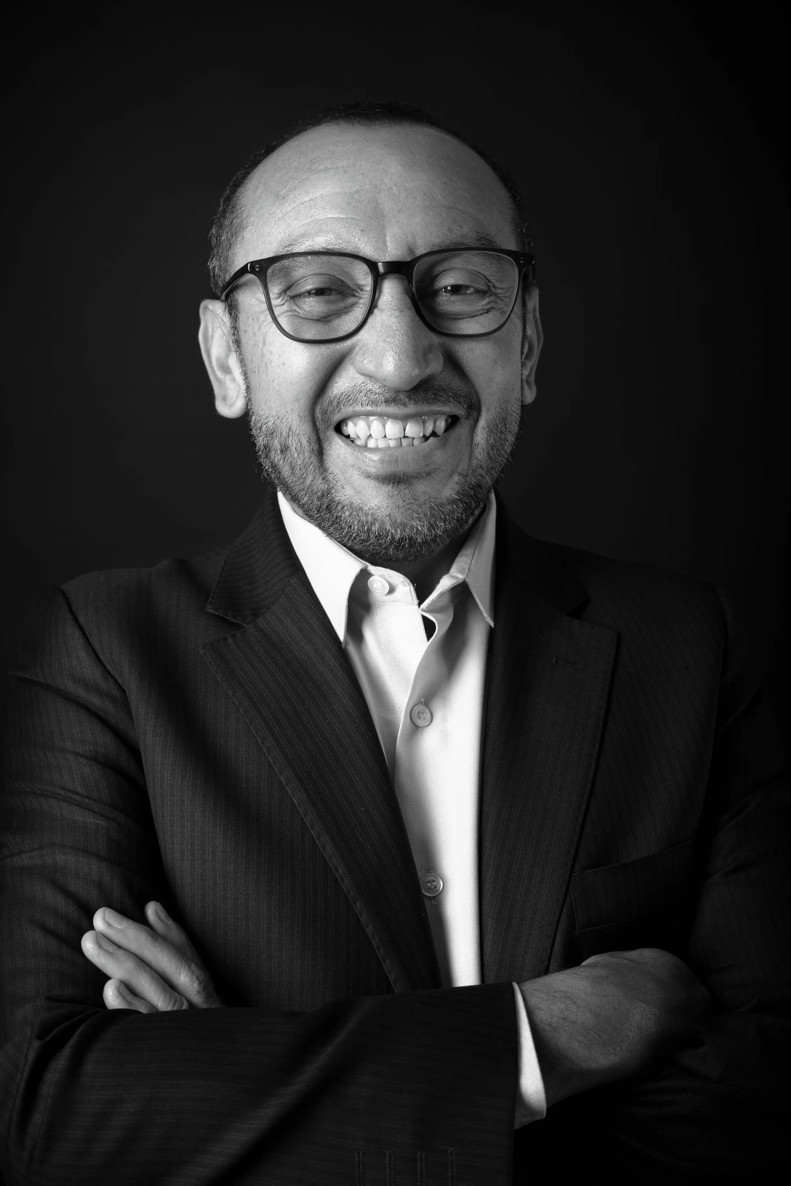 Smiling man with glasses wearing a dark suit jacket and white shirt, arms crossed against a black background.