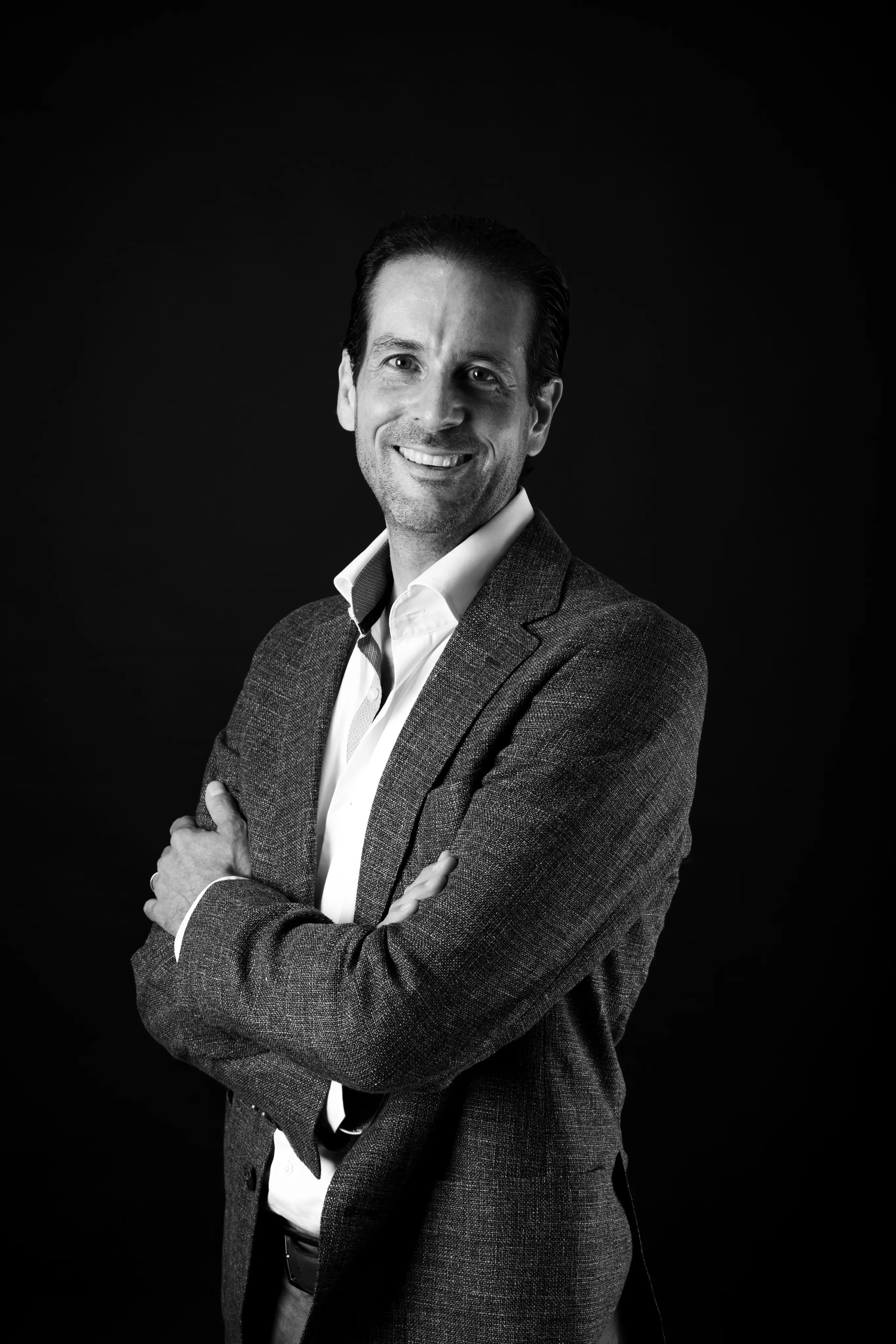Smiling man in a gray blazer and white shirt standing with arms crossed against a black background.