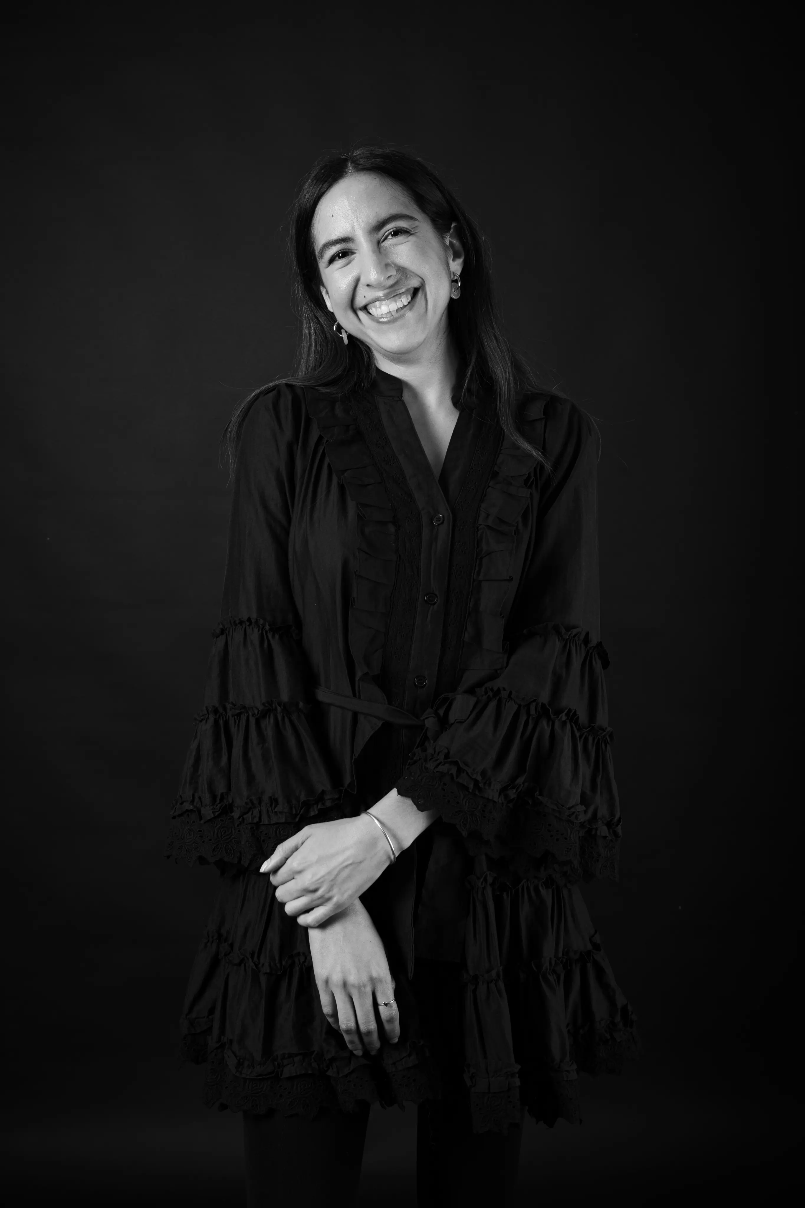 Smiling woman with long dark hair wearing a ruffled, long-sleeve dress standing against a black background.