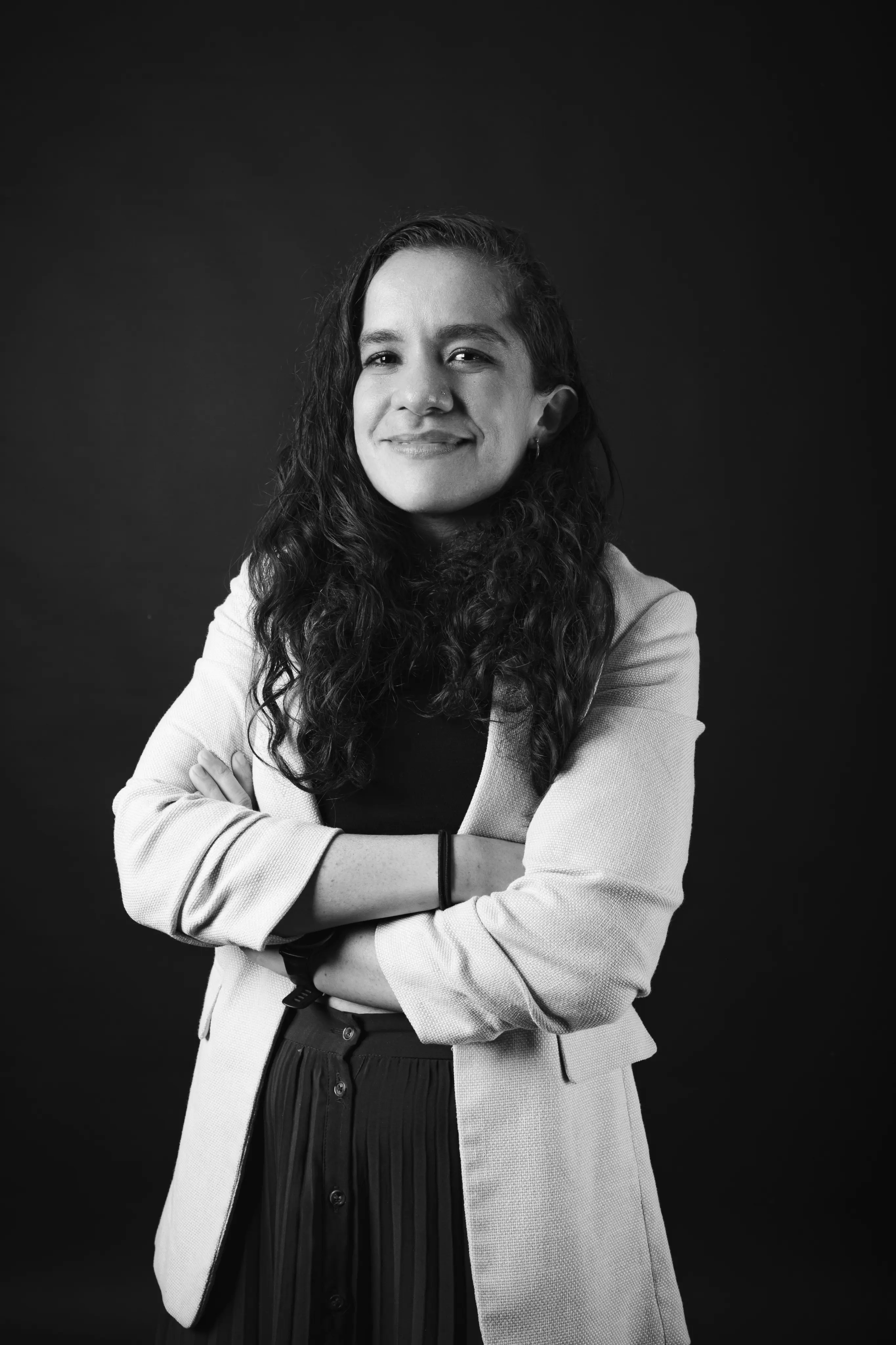 Smiling woman with long curly hair wearing a light blazer and dark top, standing with arms crossed against a black background.