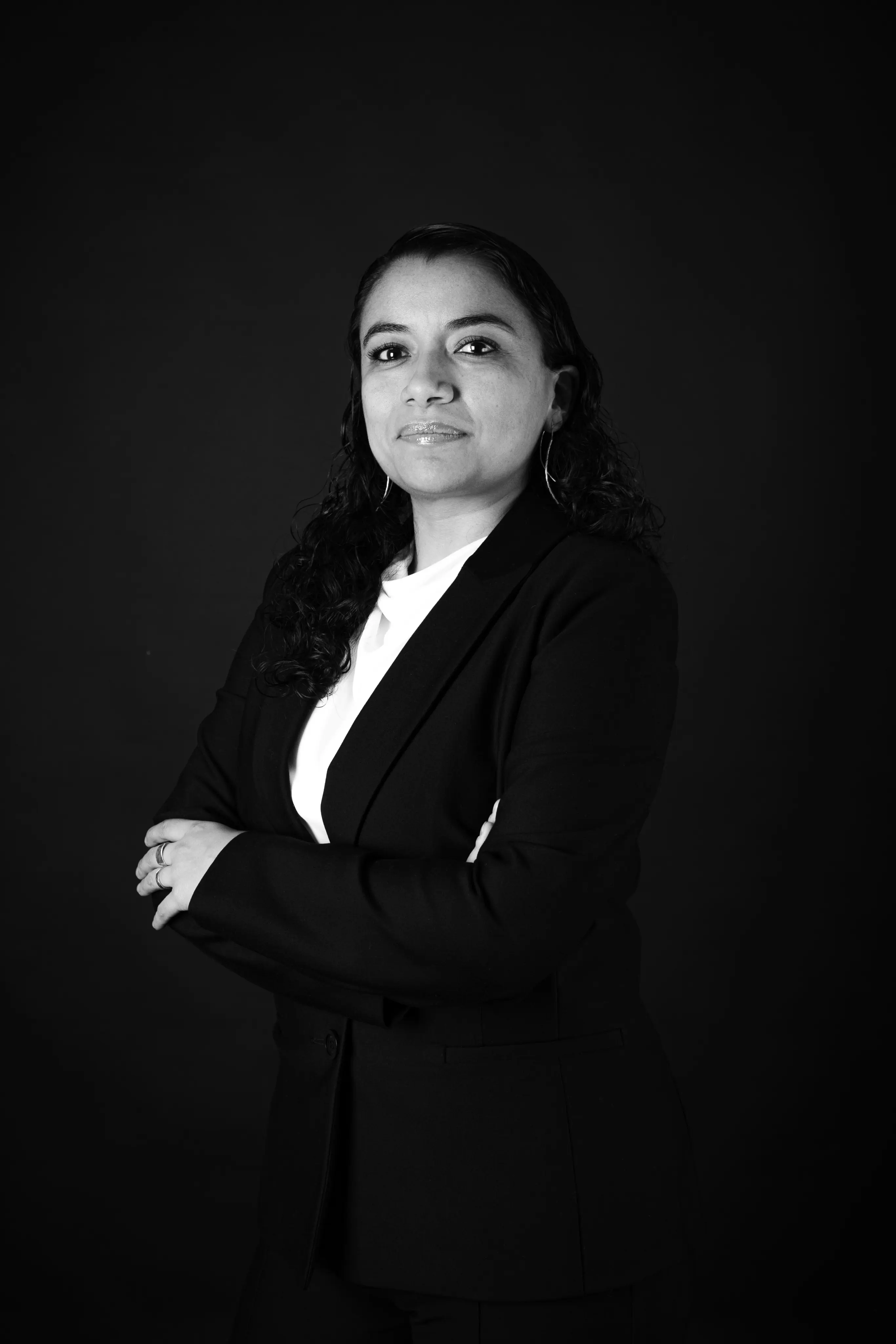 Confident woman with long dark curly hair wearing a black blazer and white shirt, standing with arms crossed against a black background.