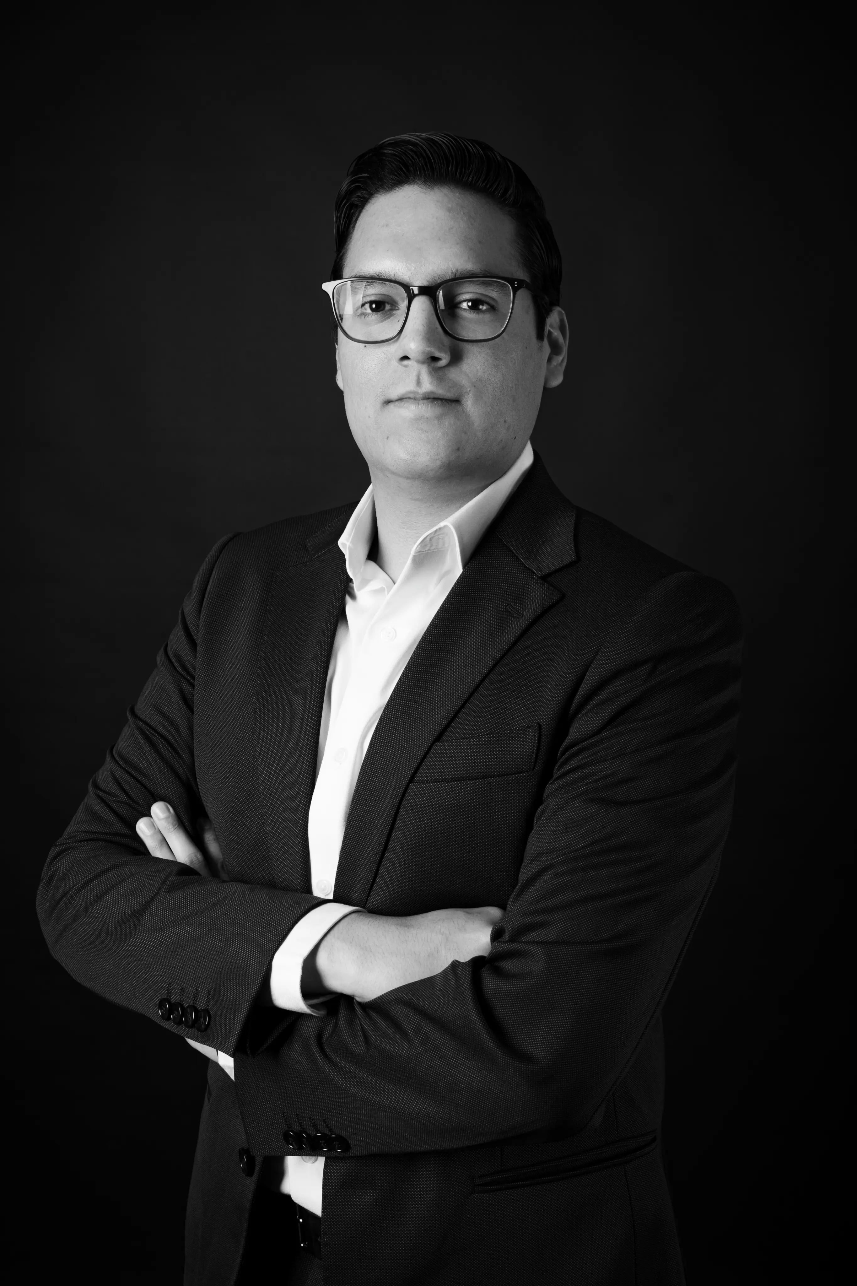 Confident man in glasses wearing a dark suit and white shirt with arms crossed against a black background.