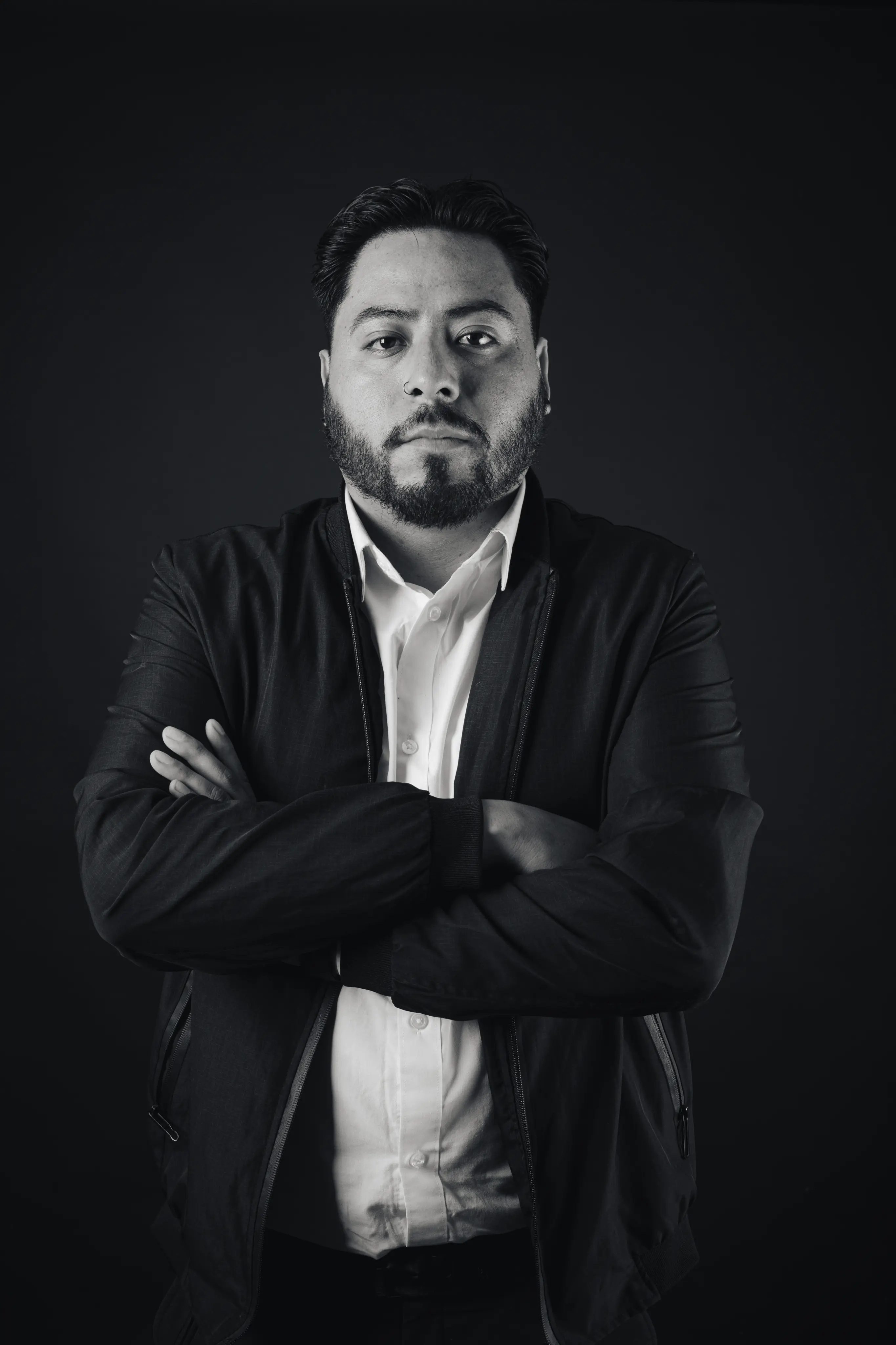 Bearded man with folded arms wearing a white shirt and dark jacket against a black background.