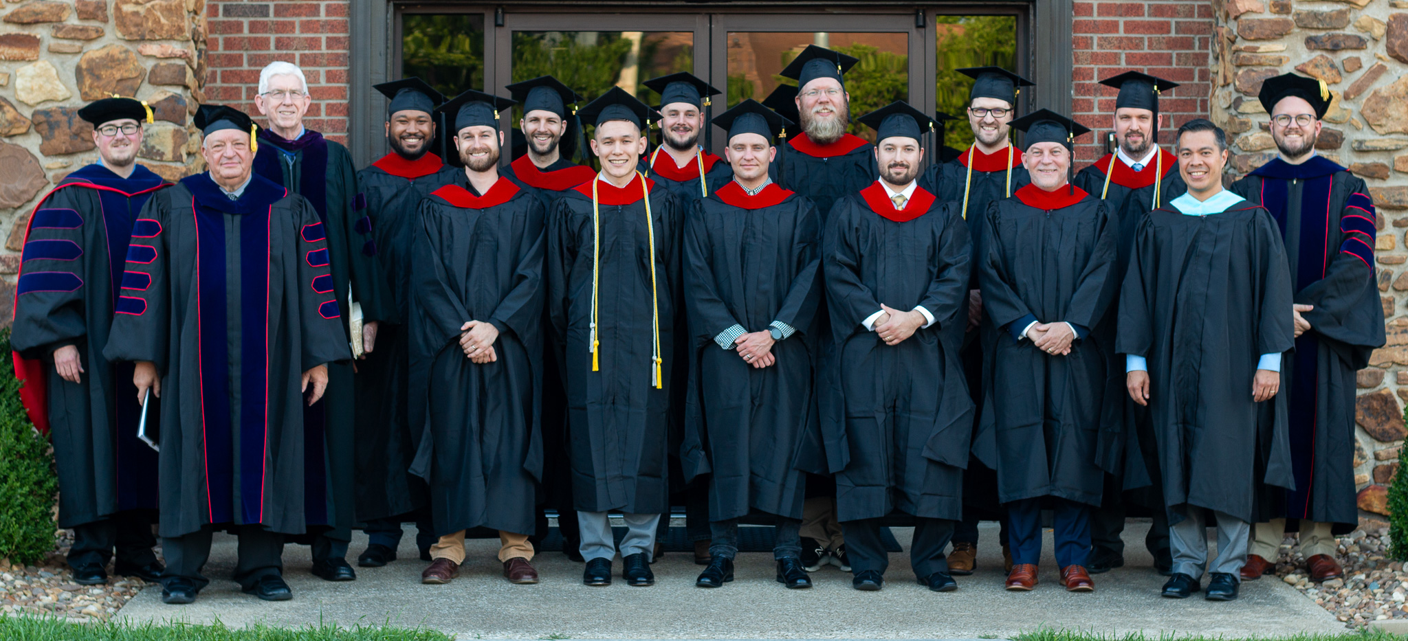 Group of graduates and faculty in academic gowns posing in front of a brick and stone building entrance.