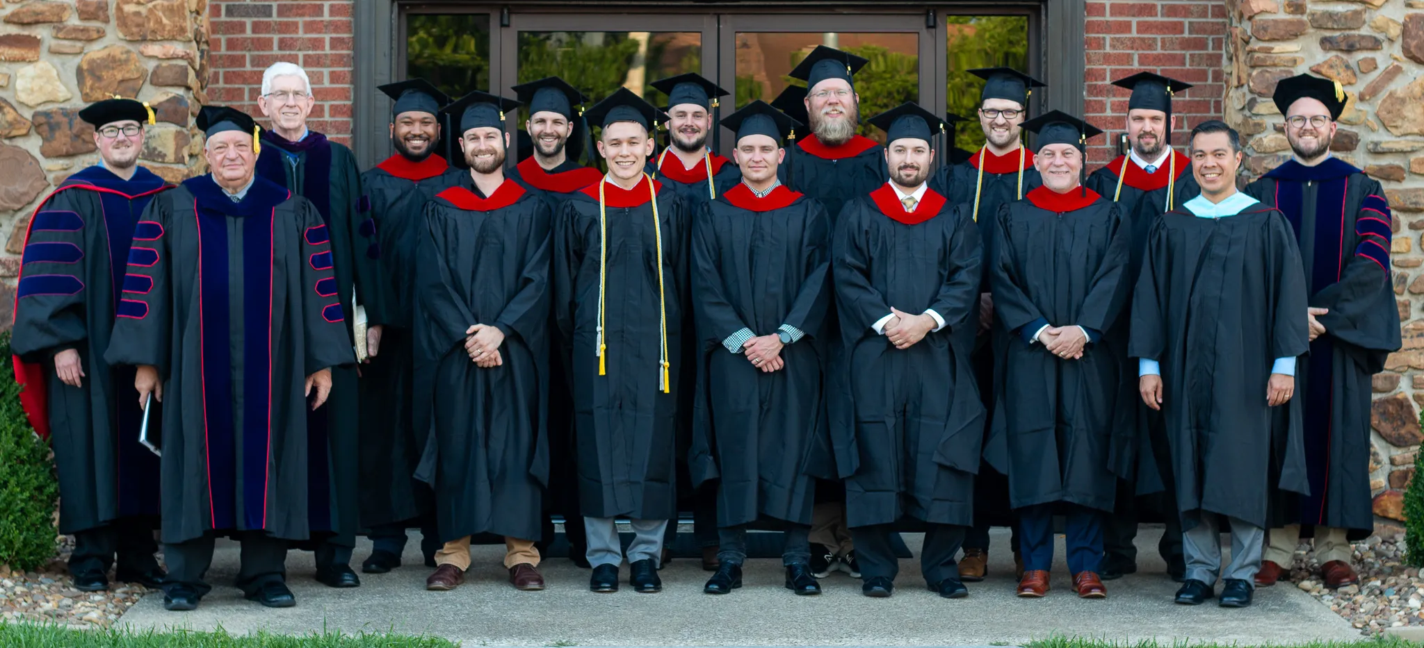 Group of graduates and faculty in academic gowns posing in front of a brick and stone building entrance.