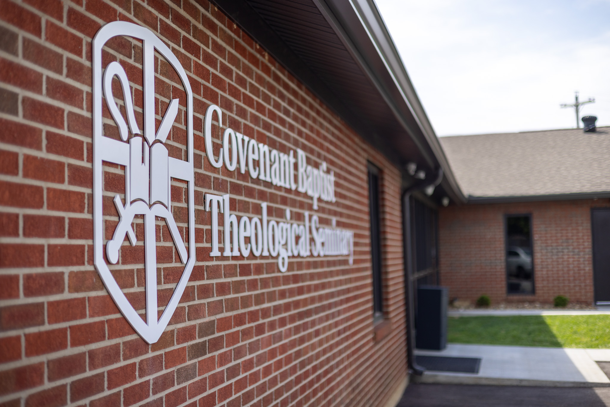 White sign with shield emblem and text 'Covenant Baptist Theological Seminary' mounted on a red brick building exterior.
