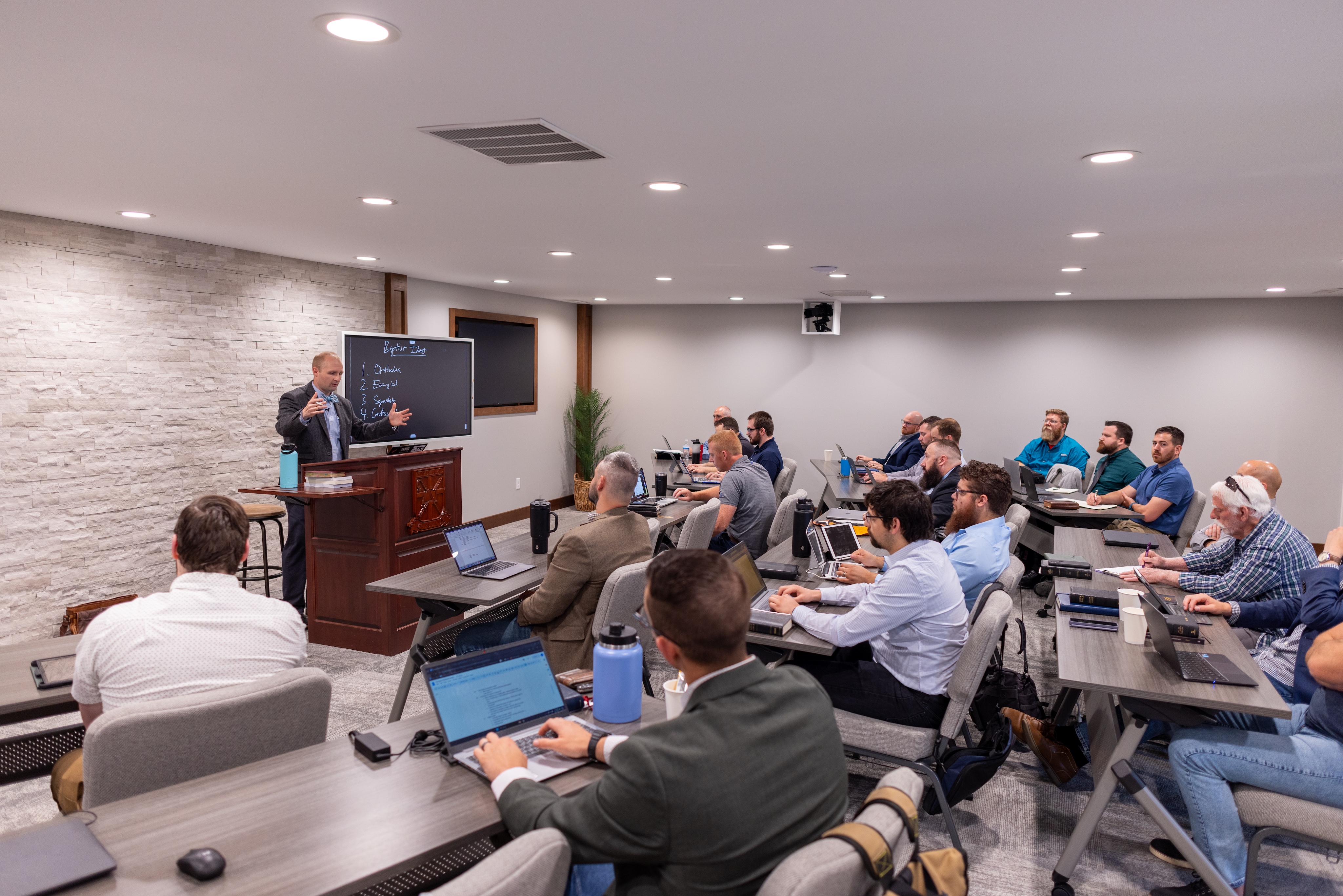 Classroom with a man standing at a lectern giving a lecture to a group of seated men working on laptops and taking notes.