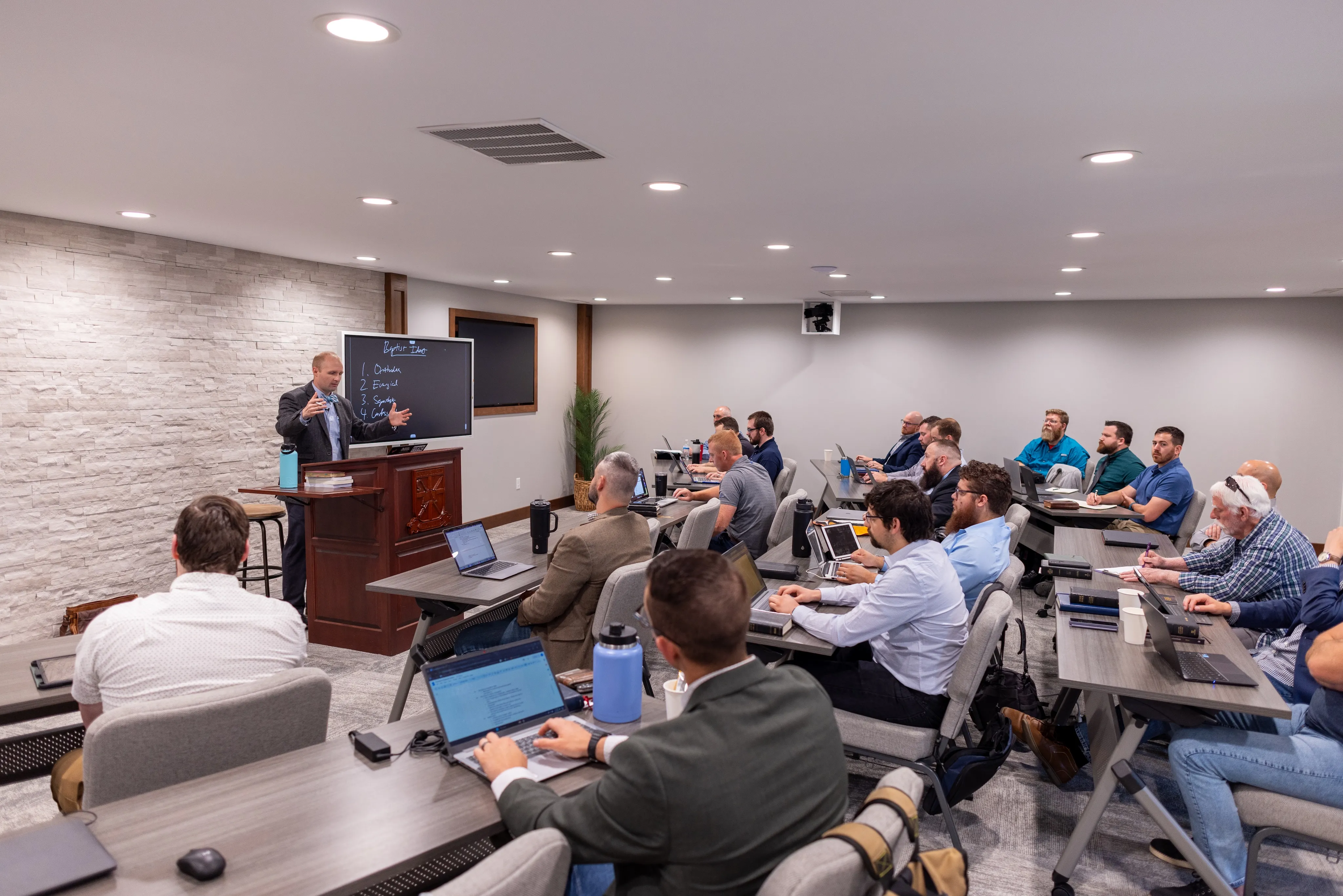 Classroom with a man standing at a lectern giving a lecture to a group of seated men working on laptops and taking notes.