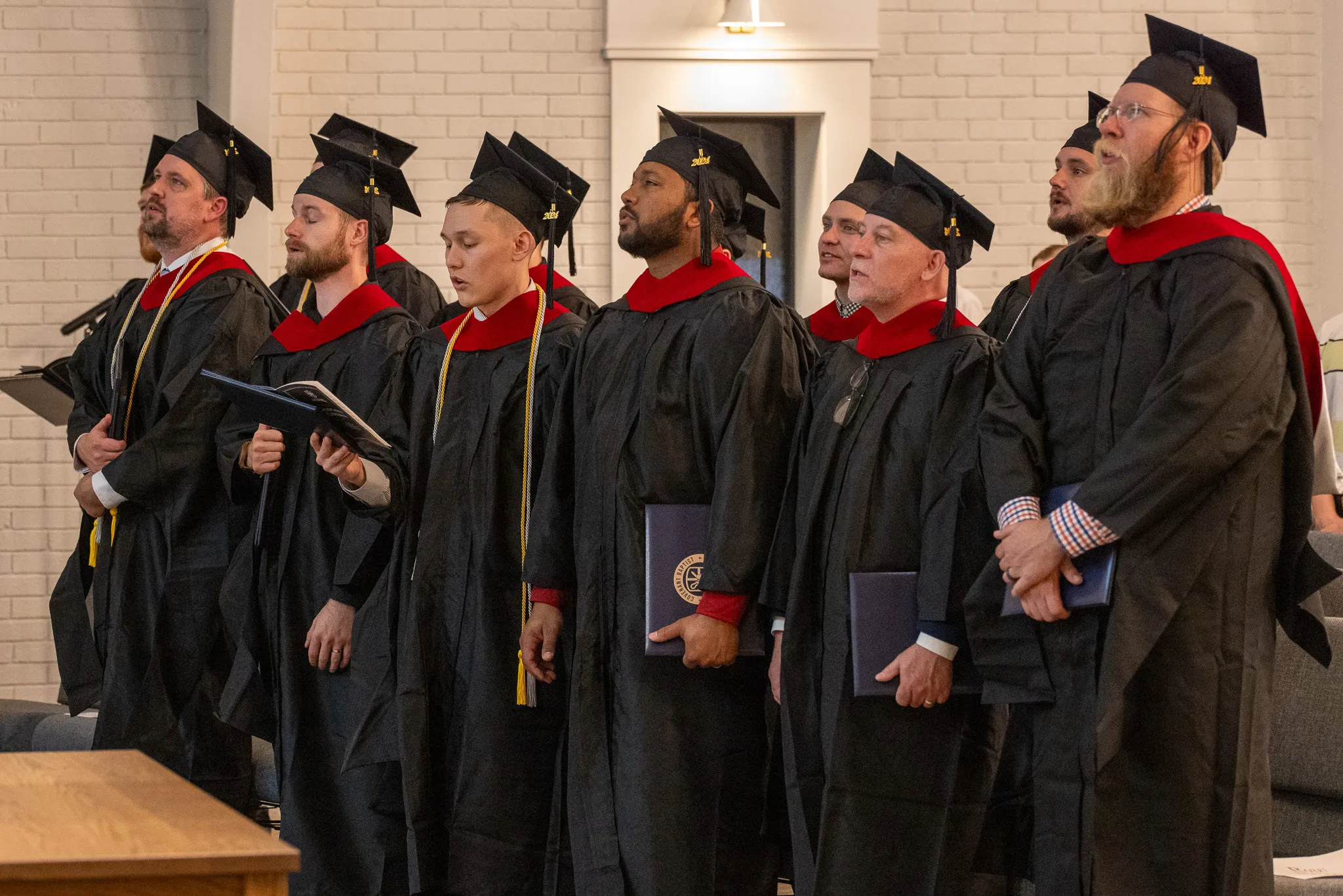 A group of male graduates in black gowns and caps with red hoods standing and holding diplomas during a ceremony.