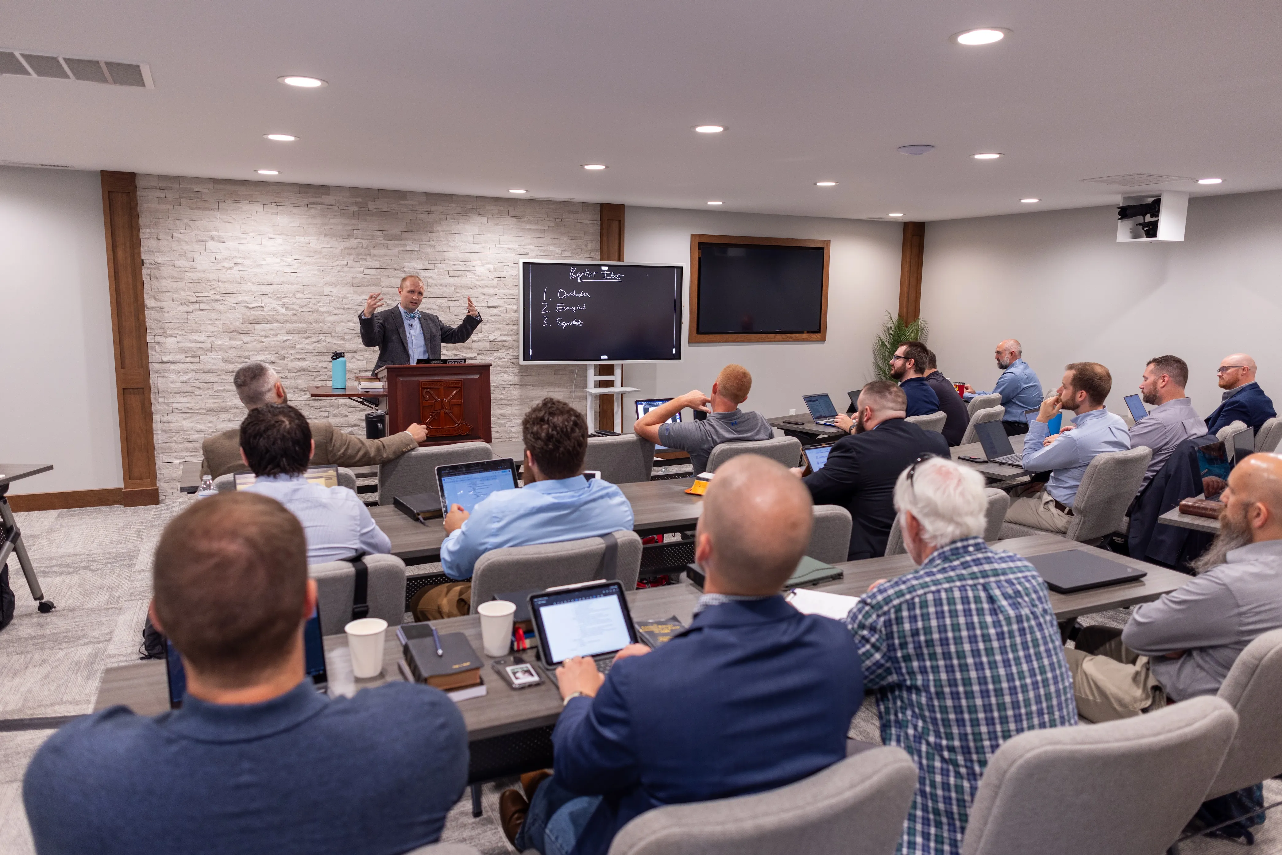 Man giving a lecture to a group of attentive men seated in a modern classroom with laptops and notebooks.