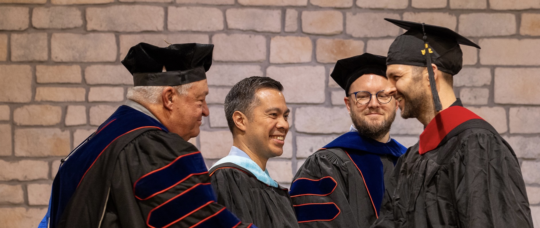Four men in academic regalia smiling at a graduation ceremony against a brick wall background.