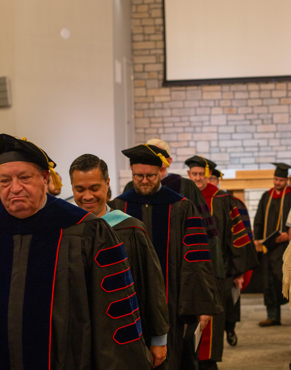 Line of faculty members wearing academic regalia and graduation caps walking indoors.