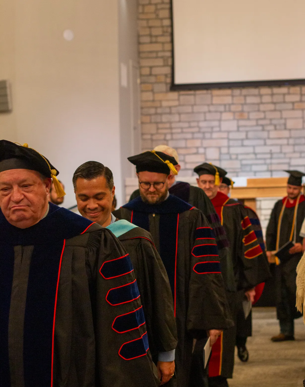 Line of faculty members wearing academic regalia and graduation caps walking indoors.