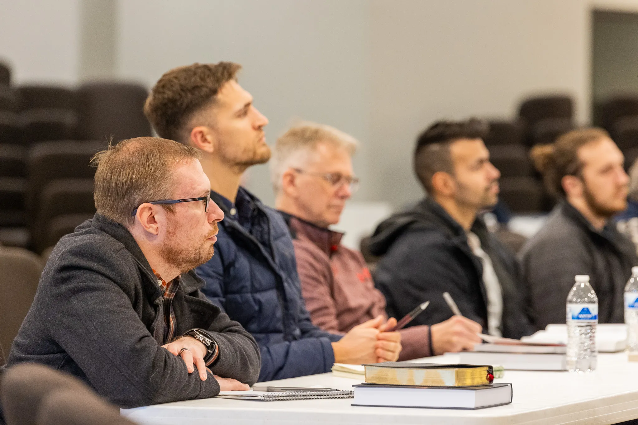Five men sitting at a table attentively listening in a classroom or seminar setting with notebooks and water bottles in front of them.