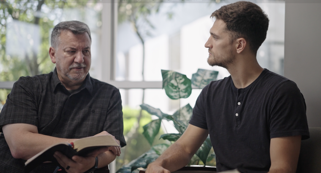 Two men sitting and having a serious conversation indoors, one holding an open book.