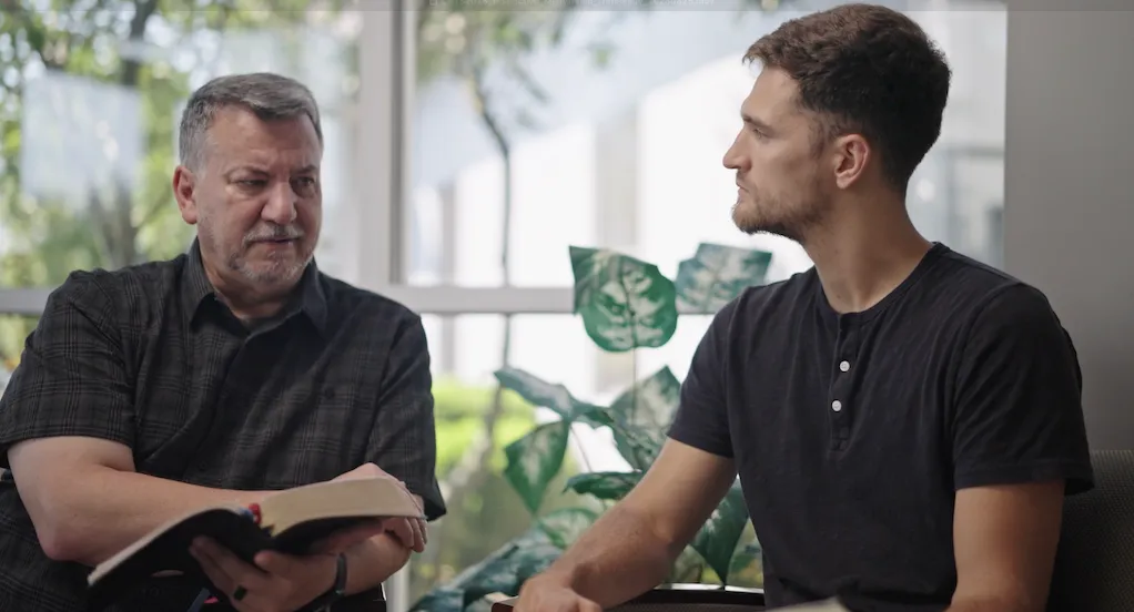 Two men sitting and having a serious conversation indoors, one holding an open book.