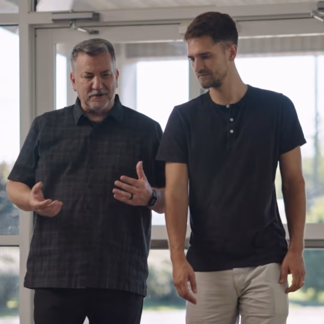 Two men walking and talking indoors near large windows, one gesturing with hands.