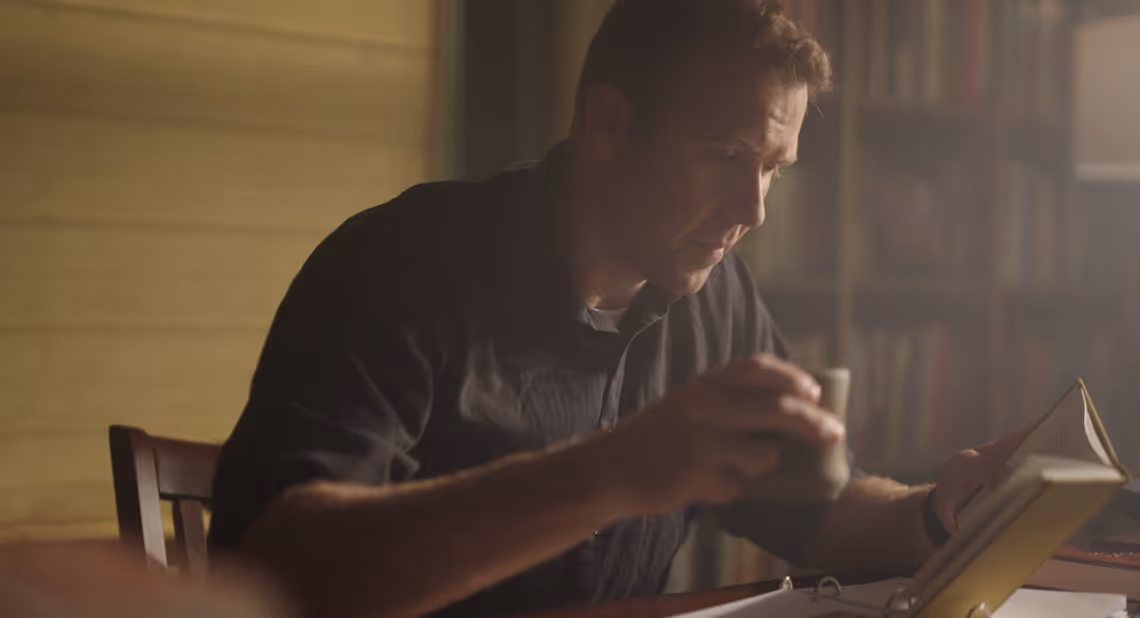 Man in a dark shirt reading a book while sitting at a table in a warmly lit room with bookshelves in the background.