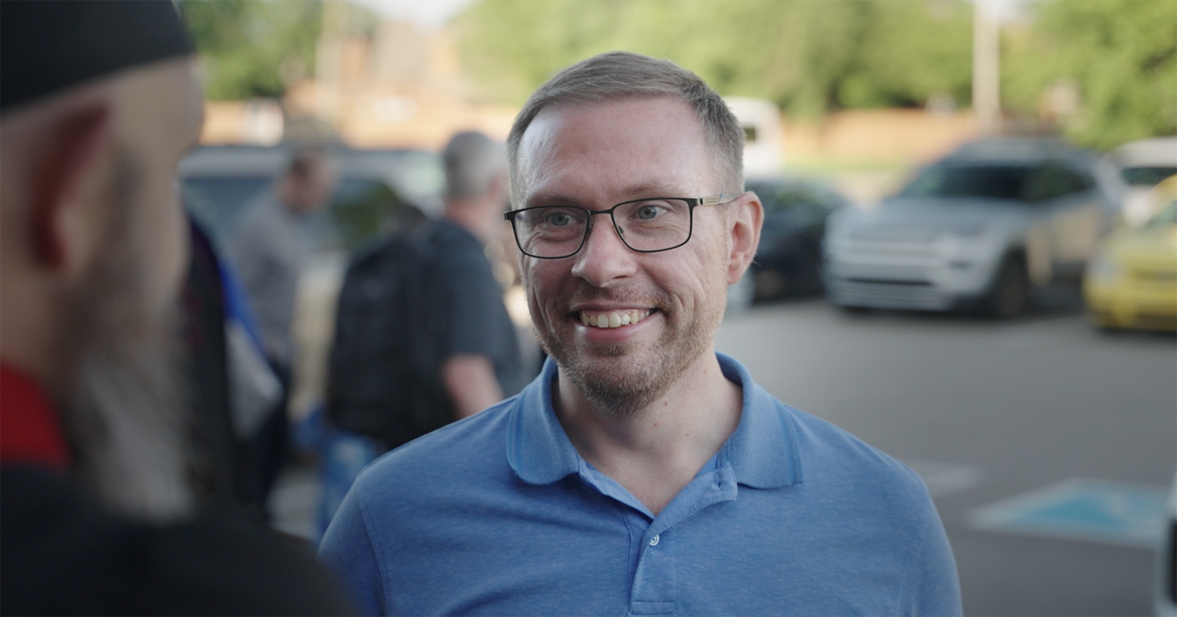 Smiling man wearing glasses and a blue polo shirt outdoors with blurred people and parked cars in the background.