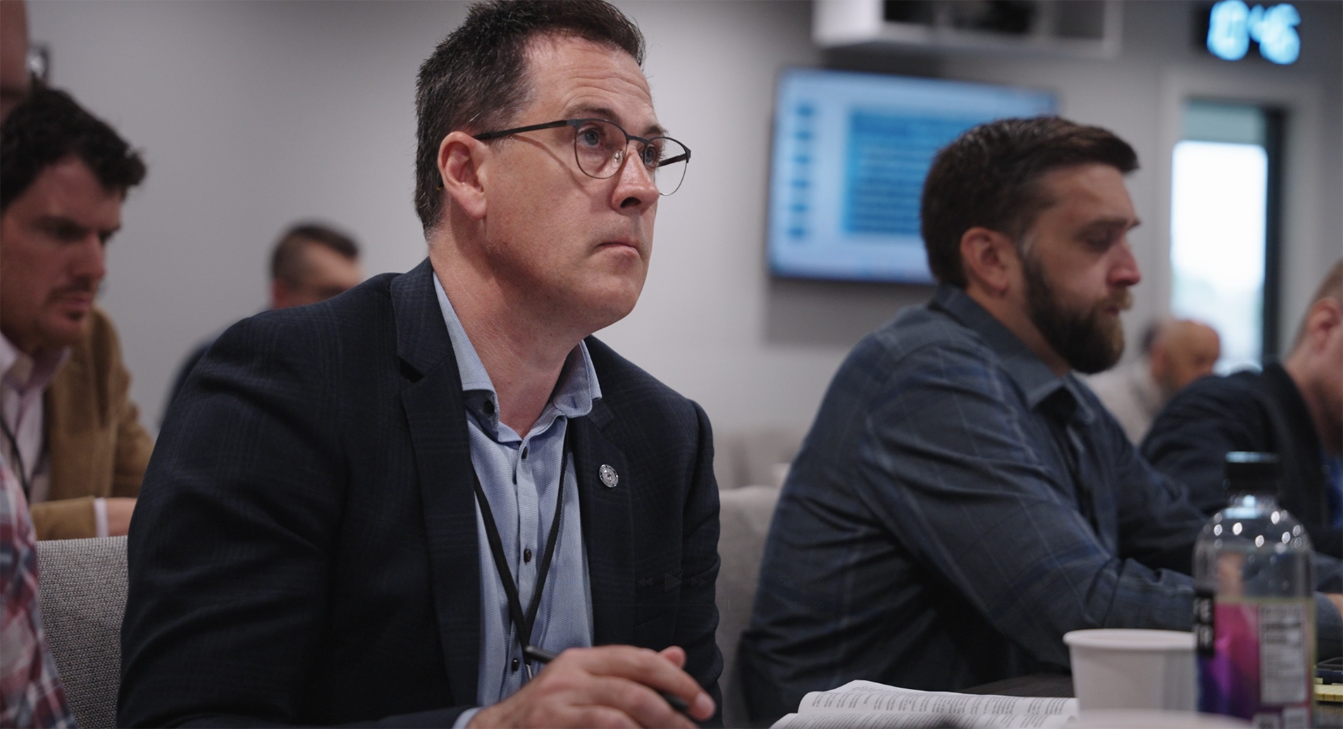 A middle-aged man wearing glasses and a suit jacket attentively listens during a meeting, with other participants sitting beside him.