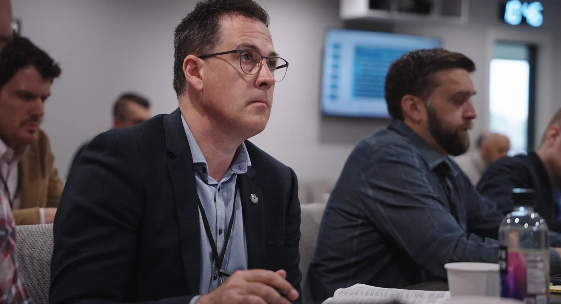 A middle-aged man wearing glasses and a suit jacket attentively listens during a meeting, with other participants sitting beside him.