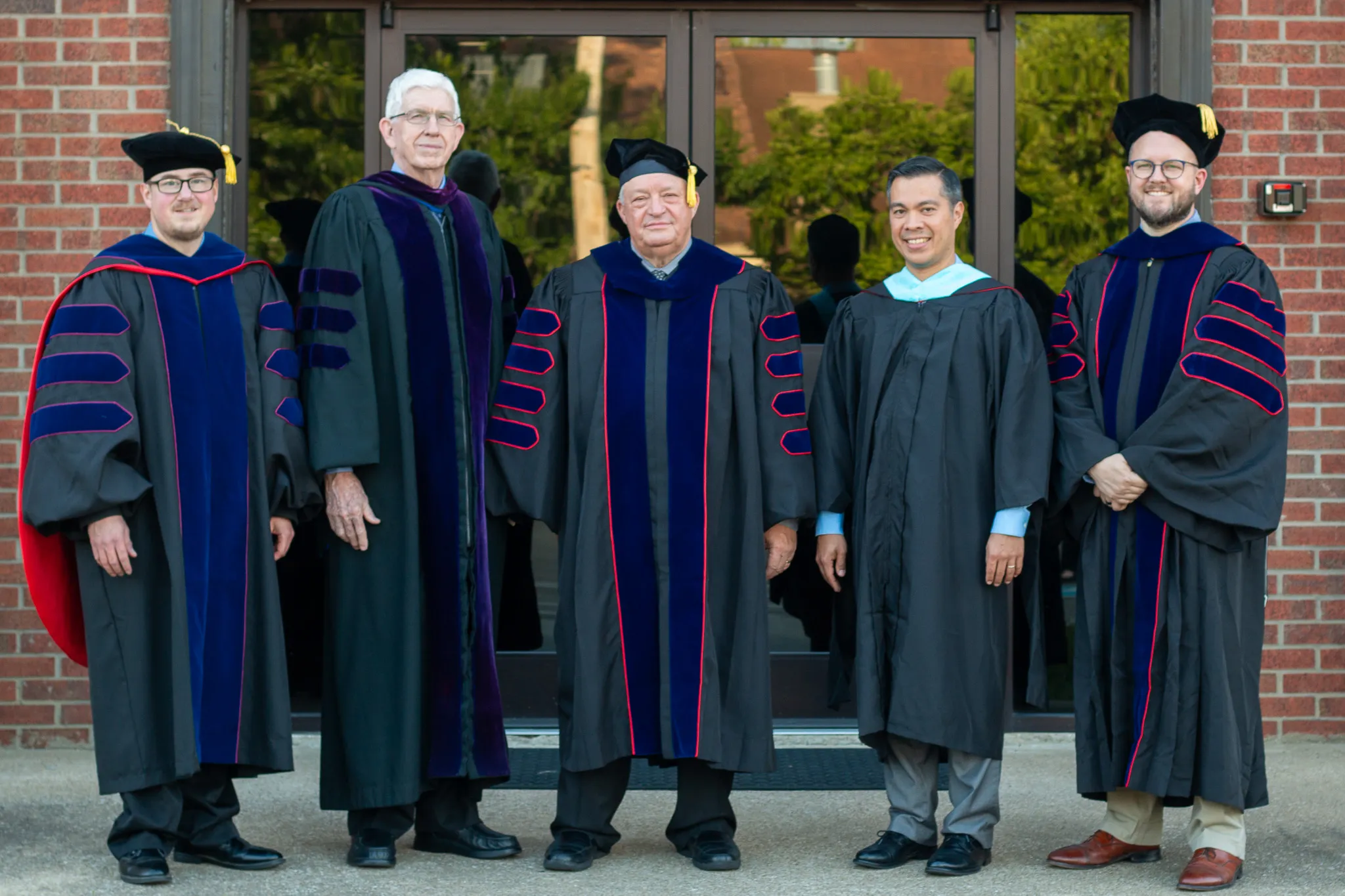 Five men standing outside a building wearing academic regalia, including caps and gowns with velvet panels and stripes.