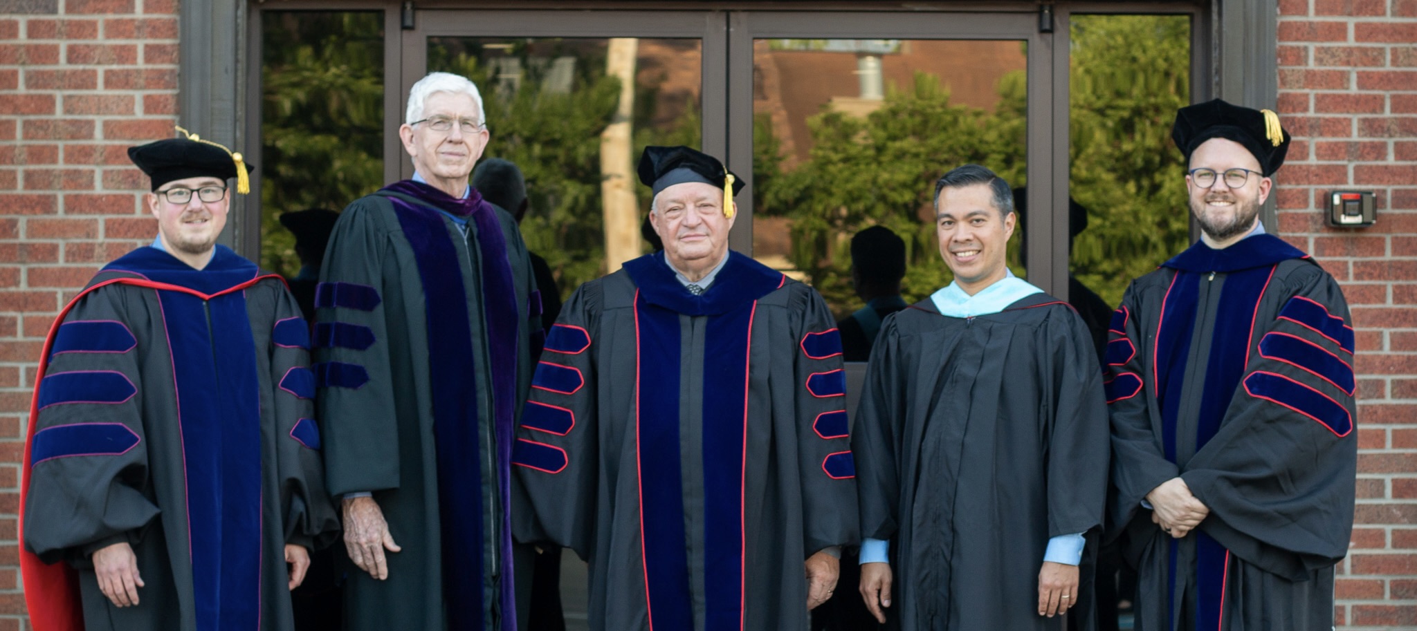 Five men in academic regalia standing in front of a glass door and brick wall, smiling at the camera.