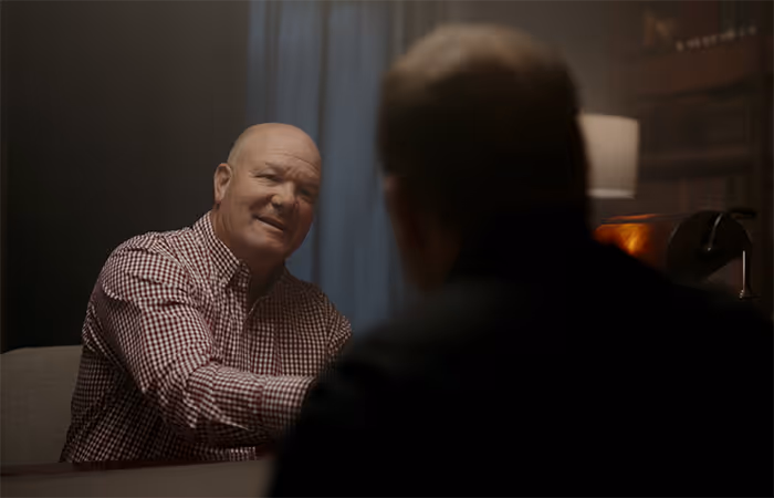 Two men sitting at a table, one facing the camera smiling and wearing a checkered shirt, the other with back to camera in a dimly lit room.
