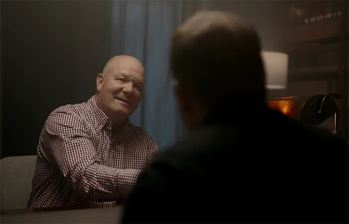 Two men sitting at a table, one facing the camera smiling and wearing a checkered shirt, the other with back to camera in a dimly lit room.