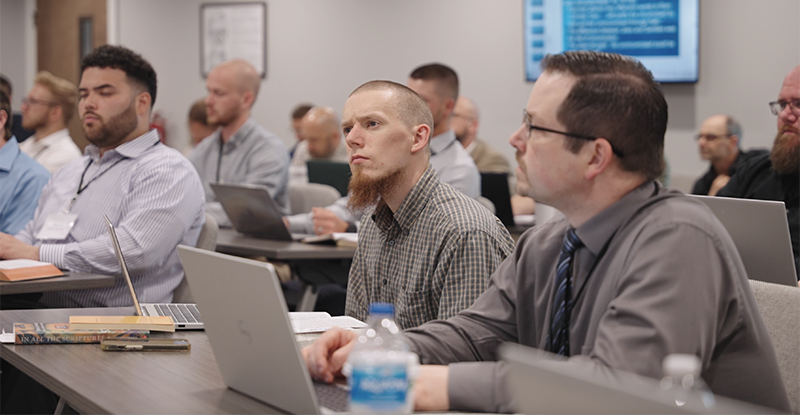 Men seated in a classroom setting, attentively listening with laptops open on desks.