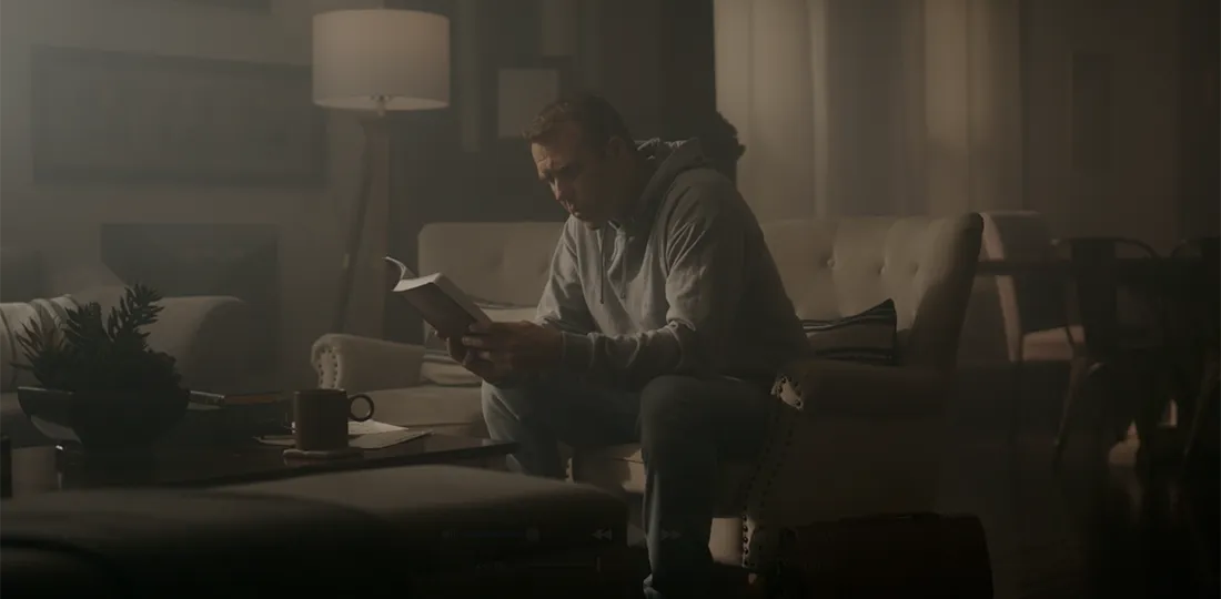 Man wearing a gray hoodie sitting on a beige armchair in a dimly lit living room, intently reading a book.